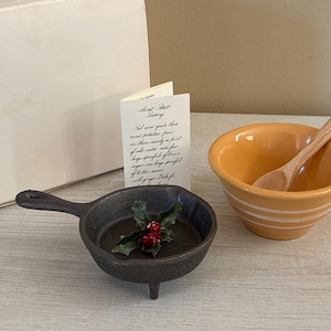 May include: A black cast iron mini skillet with three legs and a handle, containing a small holly sprig with red berries. Beside it is a yellow ceramic bowl with white stripes, a wooden spoon, a white box, and a recipe card with cursive text.