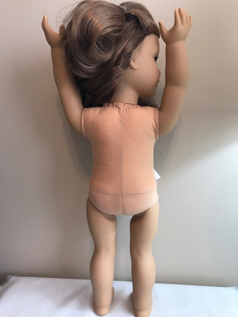 May include: A brown-haired doll wearing a nude-colored leotard with her arms raised above her head.