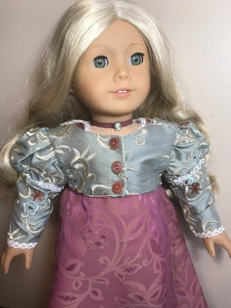 May include: A doll wearing a light blue and silver embroidered jacket with brown buttons and a pink floral skirt.
