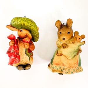 May include: Two ceramic figurines. One is a rabbit wearing a green hat and red coat, holding a bundle. The other is a mouse holding a baby mouse. Both are painted in muted tones and have a loop for hanging.