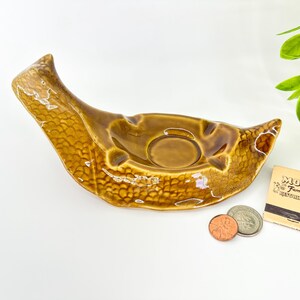 May include: A brown ceramic ashtray with a textured surface. The ashtray has a curved, elongated shape with a circular indentation. A matchbook from Mom's Family Restaurant and some coins are next to the ashtray.
