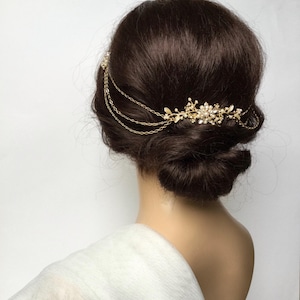 May include: A gold-coloured bridal hairpiece with floral and pearl accents. The headpiece features delicate chains and is worn on a person with dark brown hair styled in a low bun. The background is white.