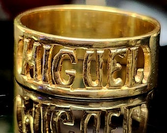 May include: A gold ring with the name "MIGUEL" cut out around the band. The ring is highly reflective, with the name appearing in a slightly darker shade of gold. The ring is displayed on a reflective surface.