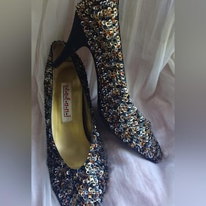 Puede incluir: Un par de zapatos de tacón con punta afilada cubiertos de lentejuelas multicolores en tonos oro, plata y negro. Los zapatos cuentan con un tacón fino y un forro interior dorado con una etiqueta blanca que dice stefani en el interior.