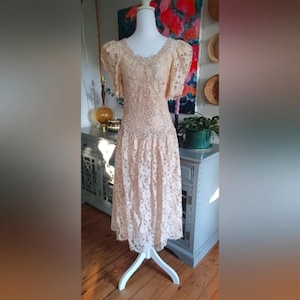 May include: A vintage peach-colored lace dress displayed on a white mannequin. The dress features a scalloped neckline, short puff sleeves, and a fitted bodice that flows into a midi-length skirt. The intricate lace detailing covers the entire dress, creating a delicate and romantic look.