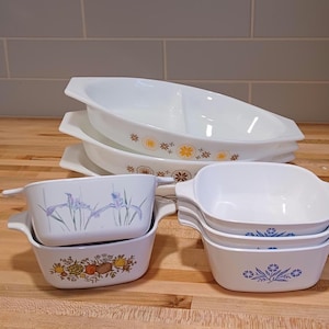 May include: A collection of vintage white CorningWare dishes. The set includes oval divided dishes with floral patterns and square casserole dishes with handles. Some dishes feature blue, yellow, and brown floral designs.