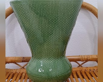 May include: A green ceramic vase with a textured, dotted pattern. The vase has a flared top, a rounded midsection, and a circular base. The vase is displayed on a woven bamboo shelf.