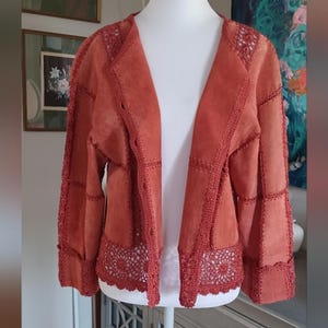 May include: A rust-colored suede jacket with crochet detailing. The open-front jacket features a patchwork design with visible stitching and crochet trim along the edges, including the collar, cuffs, and hem. The jacket is displayed on a mannequin.