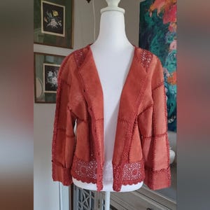 Può includere: Giacca in camoscio color ruggine con finiture all'uncinetto. La giacca presenta un design patchwork con dettagli all'uncinetto lungo i bordi, il colletto e l'orlo. La giacca aperta è esposta su un manichino.