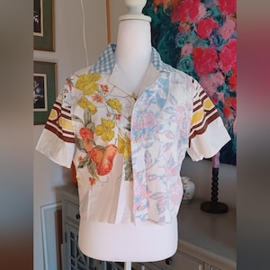 Puede incluir: Una camisa de manga corta con botones y diseño patchwork. La camisa presenta una mezcla de estampados florales y frutales en amarillo, naranja y rosa, con detalles a rayas marrones y amarillas en las mangas. El cuello es de cuadros azules y blancos.