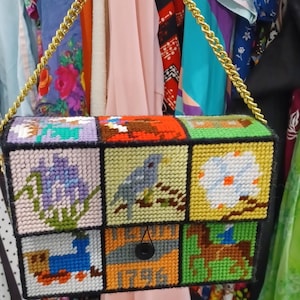 May include: A rectangular handbag crafted from plastic canvas with needlepoint designs featuring a blue bird, flowers, a train, and the text 1796. The bag has a gold-tone metal chain strap and a black button closure on the front flap.