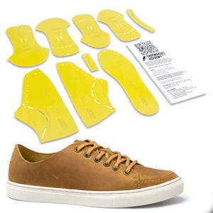 May include: A brown leather sneaker with white soles and tan laces. Yellow shoe pattern templates and a white paper with text and a QR code are also present. The templates are in various shapes and sizes.