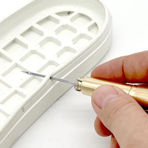 May include: A close-up of a hand using a tool to sew a white leather shoe. The tool has a brass body and a silver needle. The shoe has a grid pattern on the sole.