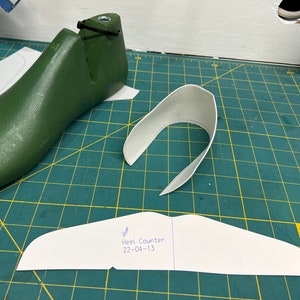 May include: A white paper pattern for a shoe heel counter, labeled "Heel Counter 22-04-13". The pattern is on a green cutting mat with a green shoe last and a white fabric piece.