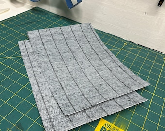 May include: Two gray and black striped fabric squares stacked on top of each other on a green cutting mat. The mat has a ruler marked in inches. A yellow label with the text "OLFA ROTARY MAT" is partially visible in the bottom right corner.