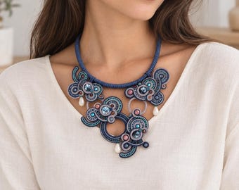 Puede incluir: Un collar llamativo con intrincado bordado de cordón de soutache azul marino, acentuado con cristales azul claro y oscuro, pequeñas cuentas redondas en tonos rosa y turquesa, y tres perlas blancas colgantes en forma de lágrima.