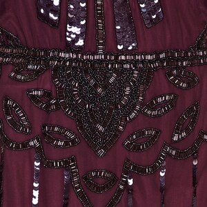 May include: Close-up of a burgundy beaded and sequined fabric. The fabric features a repeating pattern of small, dark leaves and a larger, more intricate design in the centre.
