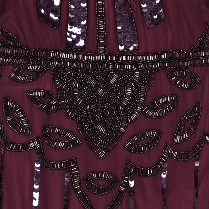 May include: Close-up of a burgundy beaded and sequined fabric. The fabric features a repeating pattern of small, dark leaves and a larger, more intricate design in the centre.