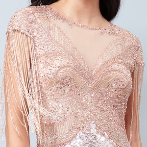 May include: A rose gold sequined dress with fringe detailing on the sleeves. The dress has a V-neckline and a fitted silhouette.
