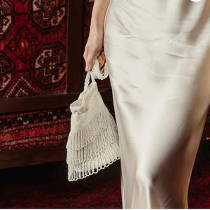 May include: A cream-colored satin slip dress paired with a matching cream-colored beaded handbag. The handbag features a braided handle and three distinct horizontal tiers of dangling beaded fringe that hang from the bottom of the bag.