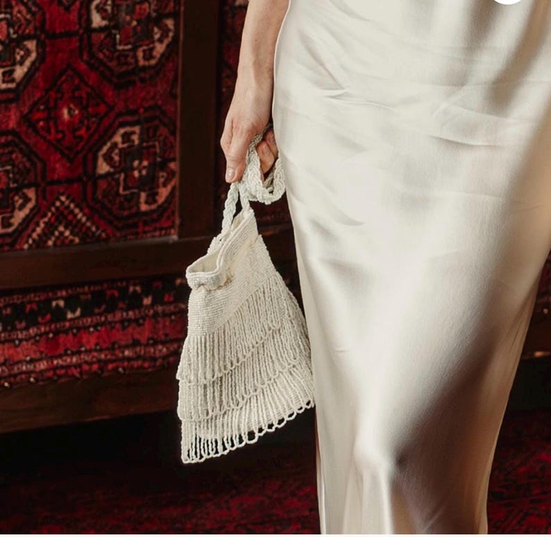 May include: A cream-colored satin slip dress paired with a matching cream-colored beaded handbag. The handbag features a braided handle and three distinct horizontal tiers of dangling beaded fringe that hang from the bottom of the bag.