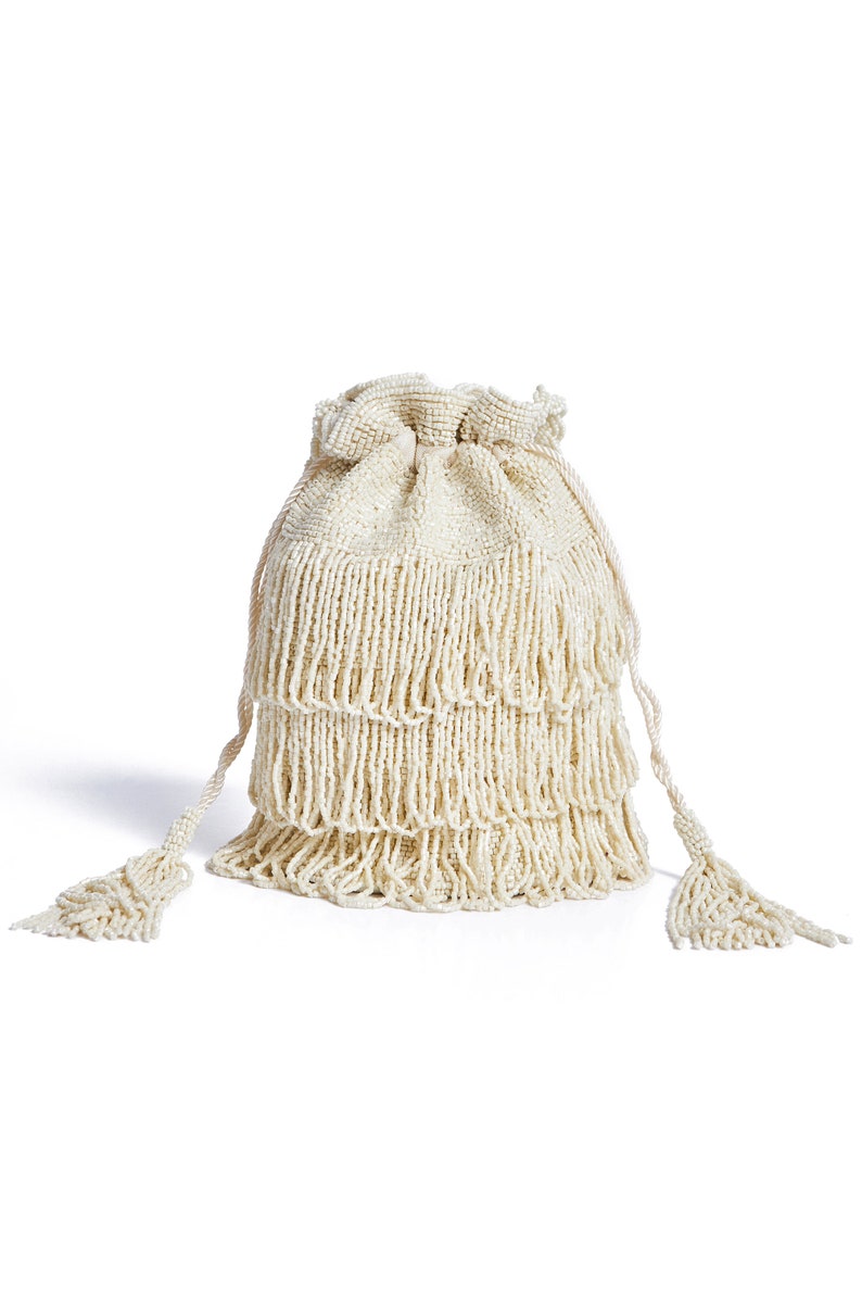 May include: A cream-colored drawstring pouch bag entirely covered in small beads. The bag features three horizontal tiers of long, dangling beaded fringe and two long braided drawstrings on each side that end in decorative beaded tassels.