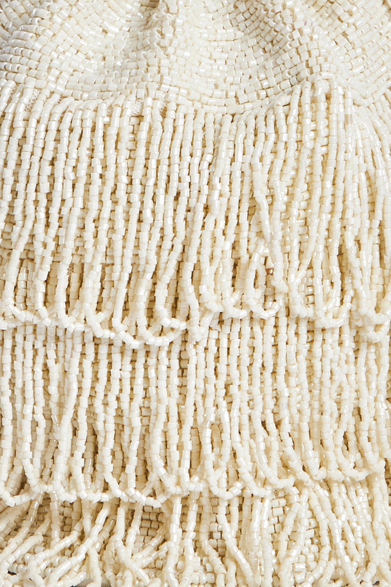 May include: A cream-colored handbag covered in layers of hanging beaded fringe. The surface is entirely composed of small, uniform, cylindrical beads arranged in vertical strands that create a textured, tiered appearance across the entire exterior.