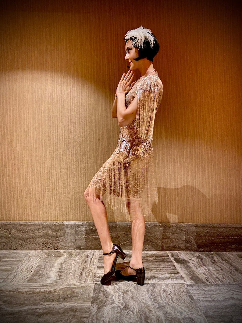May include: A woman wearing a gold sequined flapper dress with fringe, a feather headband, and black heels. She is standing in front of a brown wall.