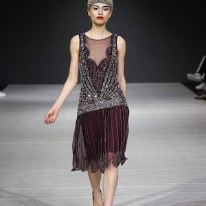 Può includere: Una modella sfila sulla passerella indossando un abito da flapper senza maniche, con paillettes, color borgogna con un'overlay trasparente. L'abito ha una scollatura con perline e un orlo a scalare. La modella indossa tacchi neri.