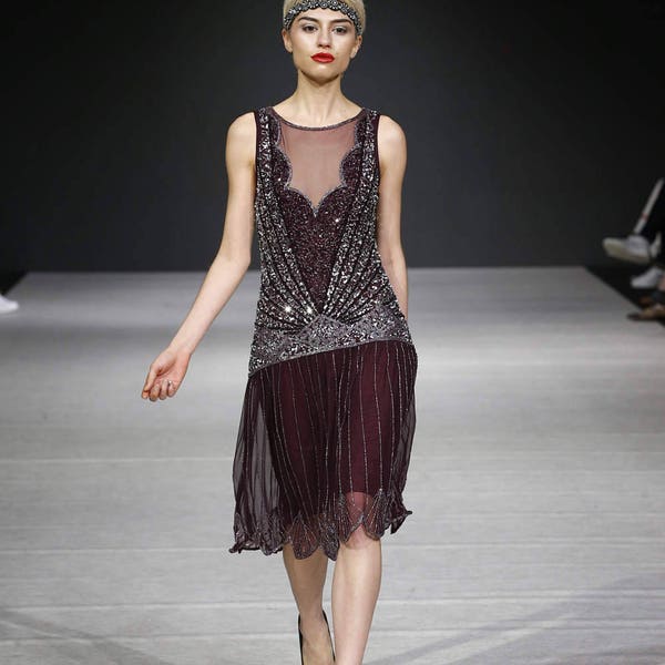 Może przedstawiać: Modelka idzie po wybiegu w bezrękawnej, cekinowej, bordowej sukience w stylu flapper z przezroczystą warstwą. Sukienka ma dekolt w kształcie litery V i opaskę na głowę z koralików.
