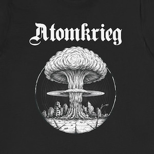 Puede incluir: Camiseta negra con la palabra "Atomkrieg" sobre un gráfico detallado de una explosión en forma de hongo. El diseño es blanco, sobre un fondo de cápsulas rojas.