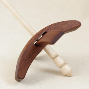May include: A drop spindle featuring a light-colored wooden shaft with a pointed, segmented tip and a dark brown, curved wooden whorl. The whorl is composed of two pieces, with one section angled outward from the central shaft of the spindle.