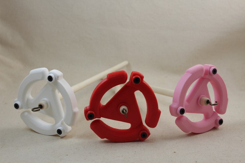 May include: Three spinning wheels with different colored bases: white, red, and pink. Each wheel has a triangular shape with three arms and a small hook in the center.