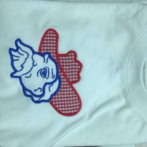 May include: White t-shirt with an embroidered red and white gingham cowboy hat and blue cartoon face.