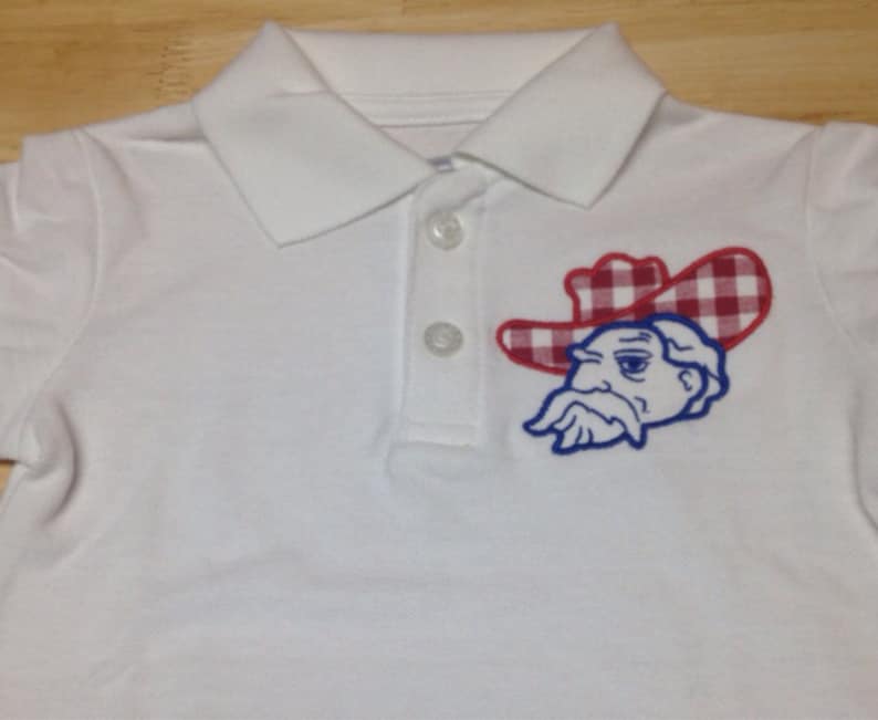 May include: White polo shirt with a blue and red embroidered logo of a cartoon character wearing a red and white checkered hat.