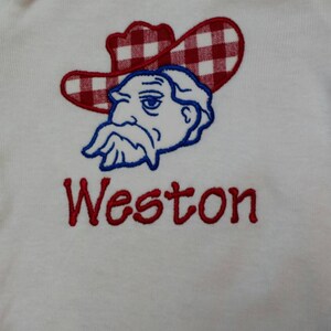 May include: White baby onesie with a red and white checkered cowboy hat and a blue cartoon face with a mustache. The name Weston is embroidered in red below the cartoon.