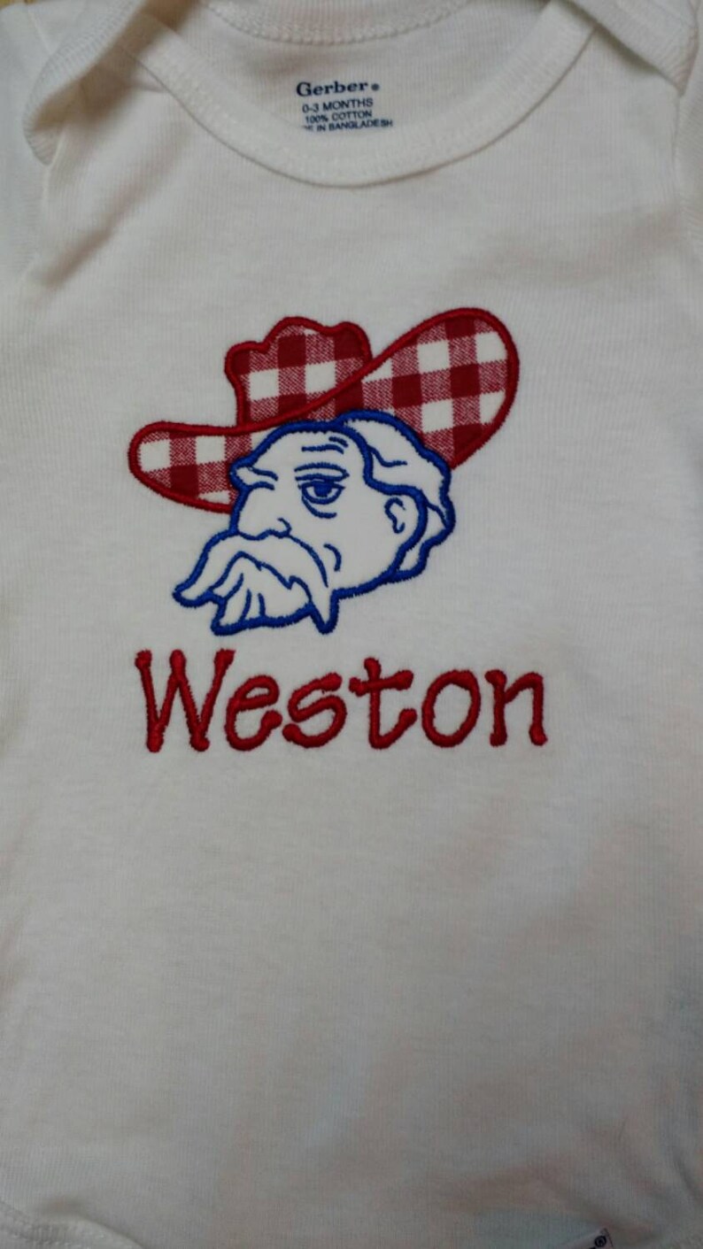 May include: White baby onesie with a red and white checkered cowboy hat and a blue cartoon face with a mustache. The name Weston is embroidered in red below the cartoon.