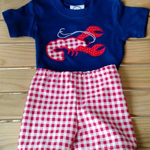 May include: A blue short-sleeved shirt with a red and white checkered lobster applique. The shirt is paired with red and white checkered shorts.