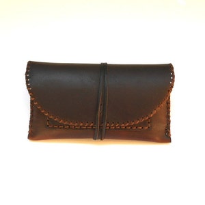 May include: A dark brown leather clutch with a flap closure and a stitched border. The clutch is secured with a leather tie. The stitching is a lighter brown color, creating a contrast against the darker leather.