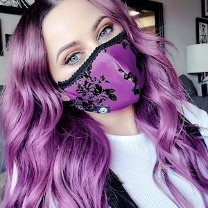 May include: A woman with long, purple hair is wearing a purple and black floral face mask. She is wearing a white shirt and black overalls.
