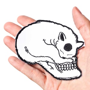 May include: White embroidered iron-on patch of a skull with a sleeping cat on top. The skull has a black outline and the cat has black eyes and a black nose.