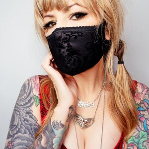 Puede incluir: Mascarilla negra con un estampado floral y ribete de encaje. La mascarilla est&aacute; hecha de una tela oscura y cubre la nariz y la boca. La persona lleva una camiseta roja y collares plateados.