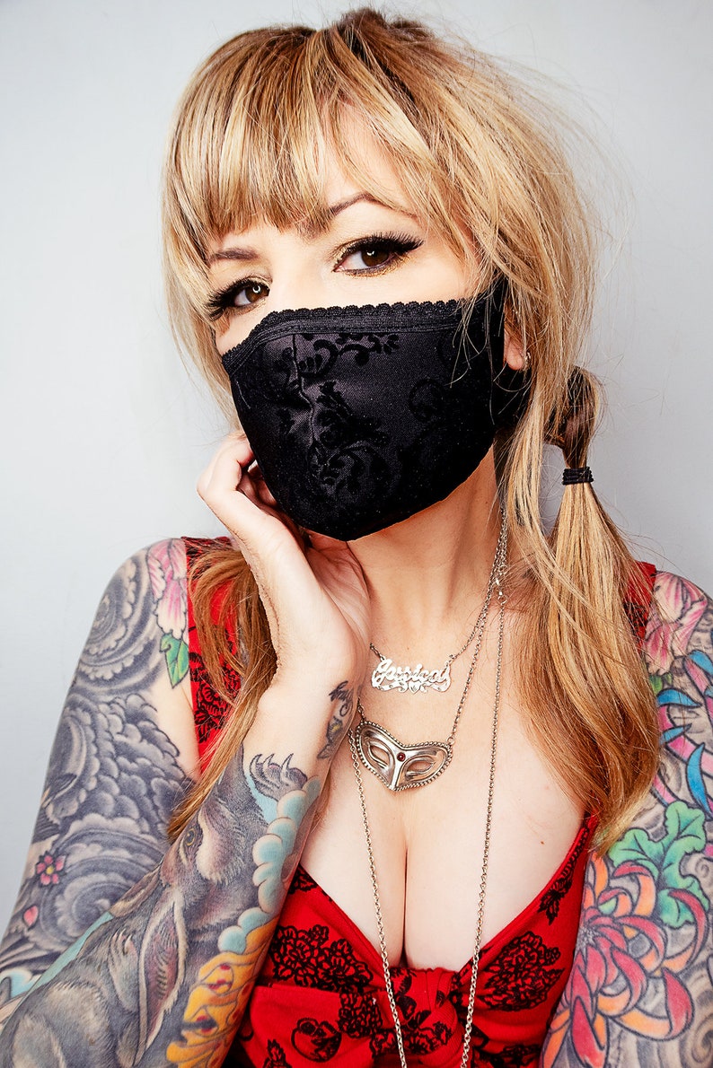 Puede incluir: Mascarilla negra con un estampado floral y ribete de encaje. La mascarilla est&aacute; hecha de una tela oscura y cubre la nariz y la boca. La persona lleva una camiseta roja y collares plateados.