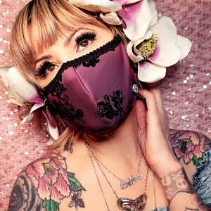 May include: A woman with a floral headband and tattoos wears a purple face mask with black lace trim. She is wearing a silver necklace with a heart pendant.