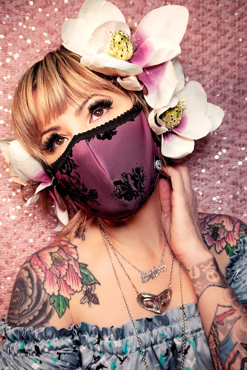 May include: A woman with a floral headband and tattoos wears a purple face mask with black lace trim. She is wearing a silver necklace with a heart pendant.