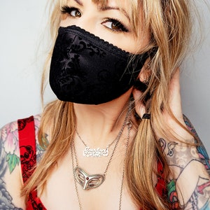 Puede incluir: Mascarilla facial de encaje negro con dise&ntilde;o floral. La mascarilla se usa con un top rojo floral y collares de plata en capas. La persona tiene cabello rubio y tatuajes visibles en los brazos.