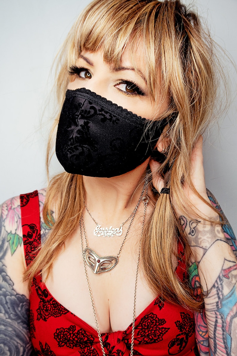 Puede incluir: Mascarilla facial de encaje negro con dise&ntilde;o floral. La mascarilla se usa con un top rojo floral y collares de plata en capas. La persona tiene cabello rubio y tatuajes visibles en los brazos.