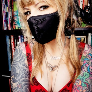 Puede incluir: Una mujer con una mascarilla negra con ribete de encaje y una blusa roja con un estampado floral negro. Tiene varios collares plateados y tatuajes en los brazos. Estanter&iacute;as en el fondo.