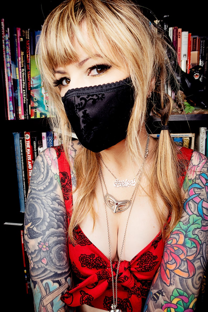 Puede incluir: Una mujer con una mascarilla negra con ribete de encaje y una blusa roja con un estampado floral negro. Tiene varios collares plateados y tatuajes en los brazos. Estanter&iacute;as en el fondo.