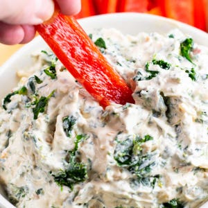 May include: A white bowl filled with creamy spinach dip. A red bell pepper strip is being dipped into the dip. The dip has visible spinach and a creamy texture, suggesting a party appetizer.